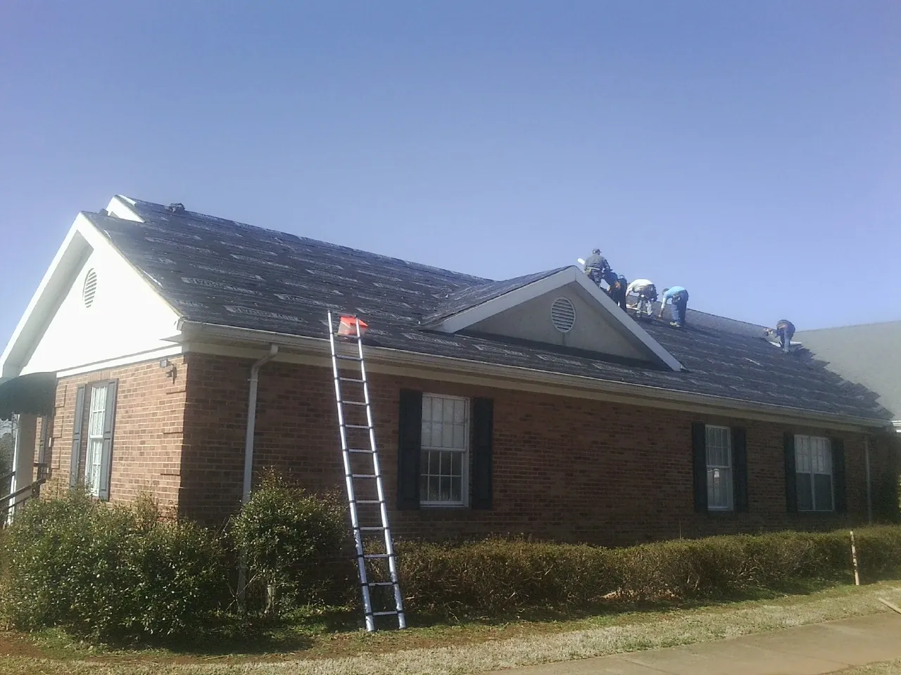 Crew tearing shingles off a brick ranch roof with a ladder set — Emergency Roof Repair work by our crew in Ionia