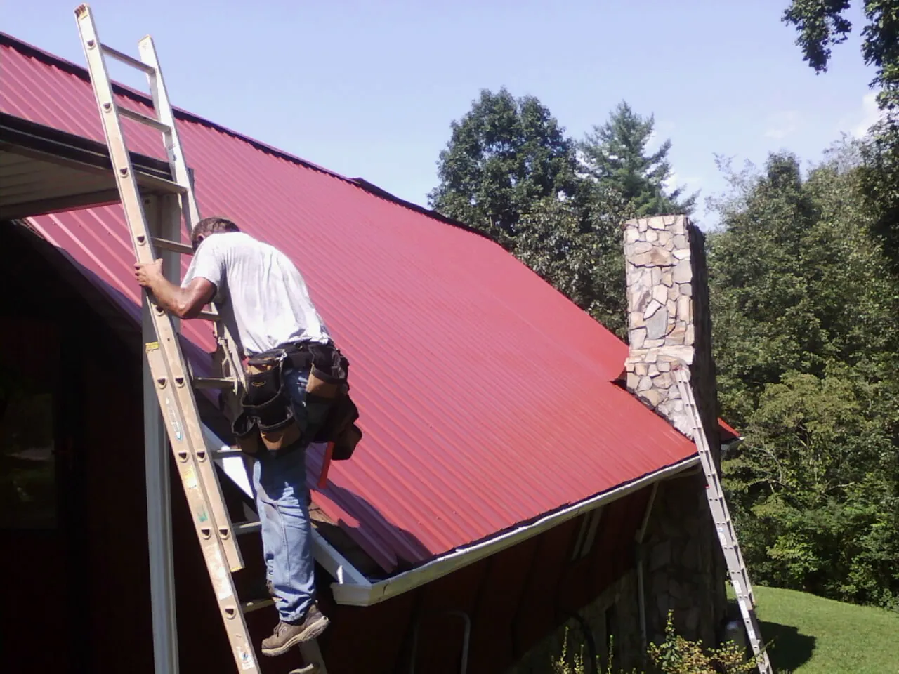 Roofer on a ladder beside a red metal roof and stone chimney — Soffit & Fascia Repair by Roofing Brothers in Ionia